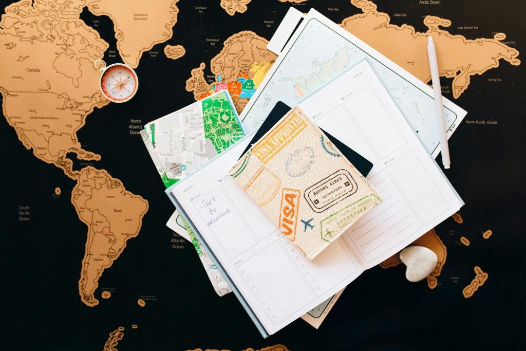 A flat lay of travel essentials, including a world map, a compass, a notebook with a handwritten note that says "Start the adventure," maps, and a passport holder with visa stamps, representing the planning phase of a global adventure or journey.